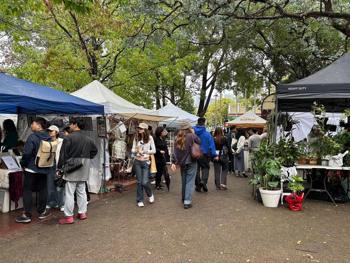 Multitudes curiosean entre coloridos puestos de mercado bajo frondosos árboles en Glebe Markets, con plantas en maceta, artículos artesanales y ropa vintage en exhibición.