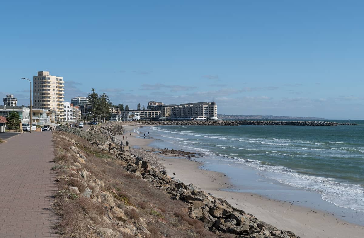 View of Glenelg Beach with rocky shoreline, sandy beach, and the foreshore lined with hotels and apartments under a clear blue sky.