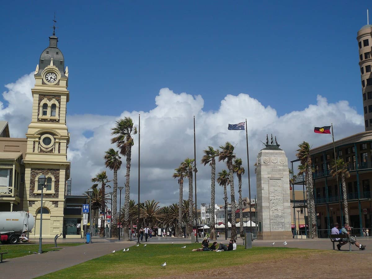 หอนาฬิกาของศาลาว่าการเมืองประวัติศาสตร์ของ Glenelg อนุสรณ์สงคราม ต้นปาล์ม และธงชาติ ใต้ท้องฟ้าสีฟ้าสดใสที่มีเมฆกระจาย