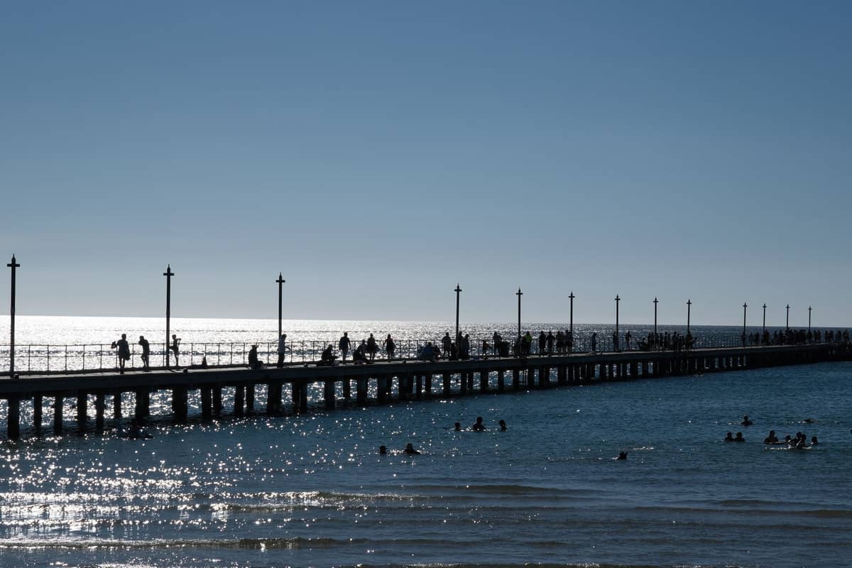 Wide view of Glenelg Jetty stretching over the water, with people walking and swimming around, sunlight sparkling on the sea below.