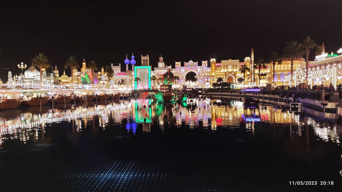 Nachtansicht des Global Village Dubai mit beleuchteten Länderpavillons, bunten Lichtern, die sich im Wasser spiegeln, und festlichen Menschenmassen an der Uferpromenade.