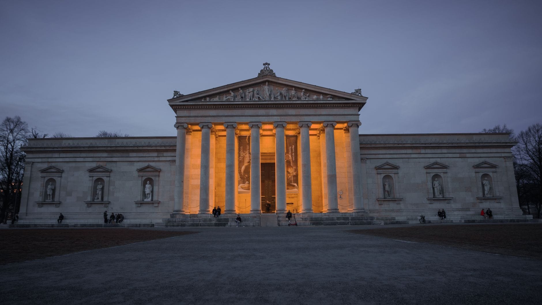 ด้านหน้าพิพิธภัณฑ์ Glyptothek ในมิวนิกช่วงหัวค่ำ สถาปัตยกรรมนีโอคลาสสิกมีเสาสูง 6 ต้น และประติมากรรมระหว่างเสา ส่องไฟเหลืองนวล ถ่ายจากมุมตรง