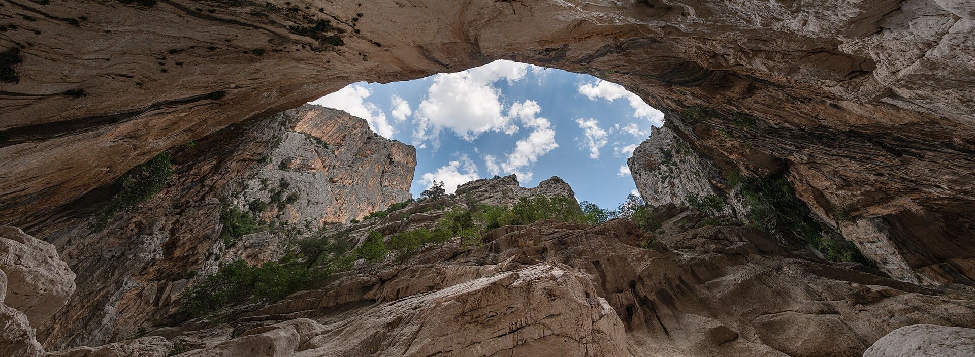 Weitwinkelansicht aus dem Inneren der Gola di Su Gorropu mit aufragenden Cañonwänden, felsigem Gelände, grünen Sträuchern und einem blauen Himmel mit vereinzelten Wolken.