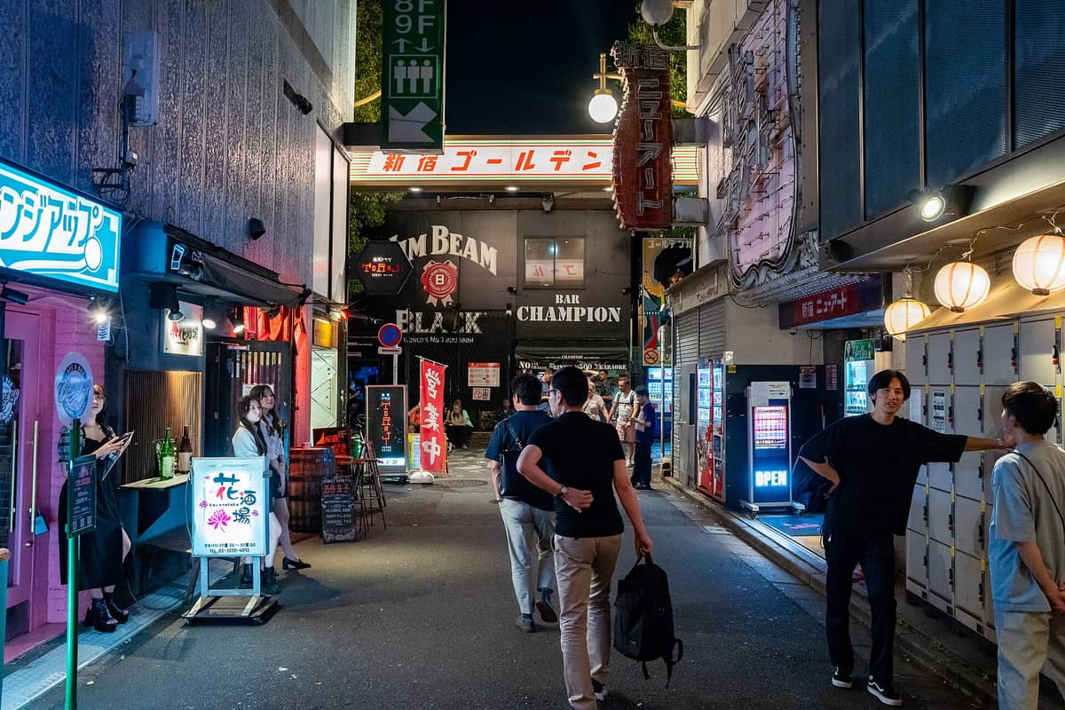 Night scene at the entrance of Golden Gai in Shinjuku, Tokyo, with glowing lanterns, small bars, and people walking the narrow alley.