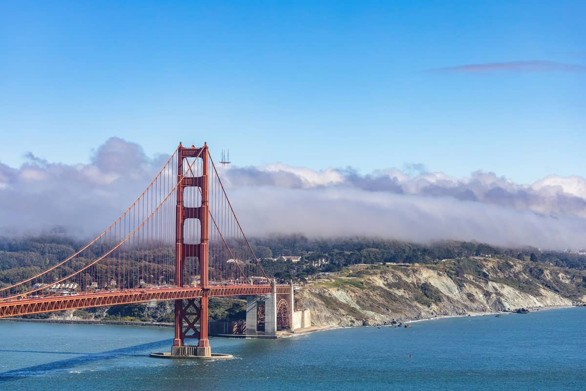 El puente Golden Gate se extiende sobre aguas azules con niebla baja y cielo despejado, mostrando sus torres rojas y las colinas del condado de Marin al fondo.
