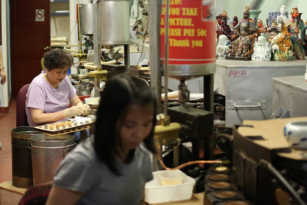 Interior de la Golden Gate Fortune Cookie Factory: una mujer dobla galletas de la fortuna a mano junto a las máquinas y bandejas de producción.