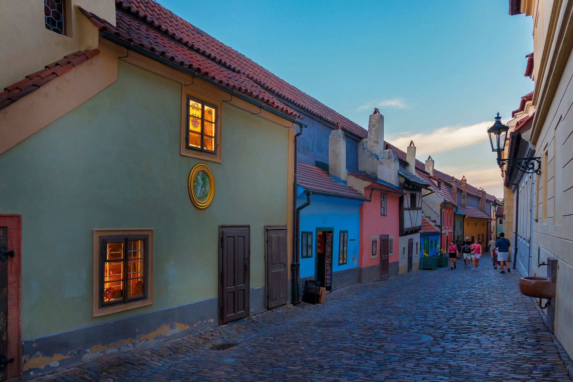Bunte, winzige historische Häuschen säumen das Kopfsteinpflaster des Goldenen Gässchens unter blauem Himmel, warmes Licht scheint aus den Fenstern und einige Touristen schlendern die Gasse entlang.