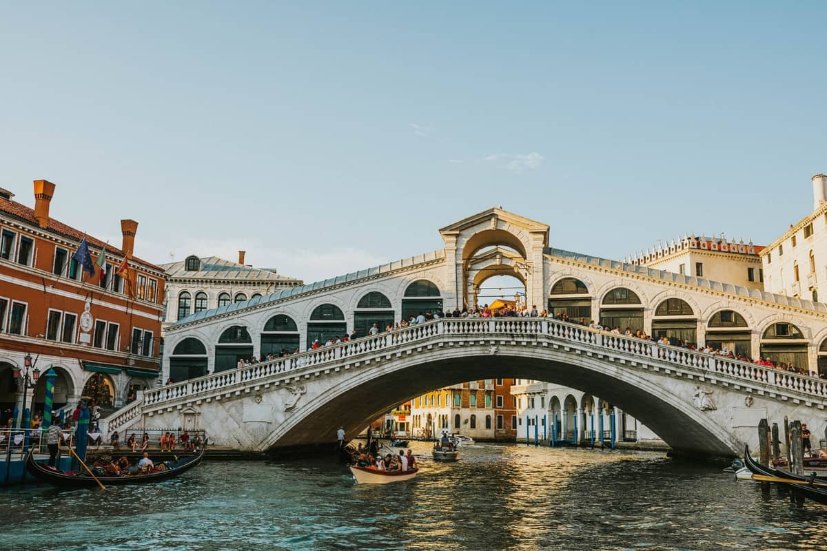 Mehrere Gondeln mit Passagieren gleiten am Nachmittag bei hellem Sonnenlicht unter der Rialtobrücke auf dem Canal Grande entlang.