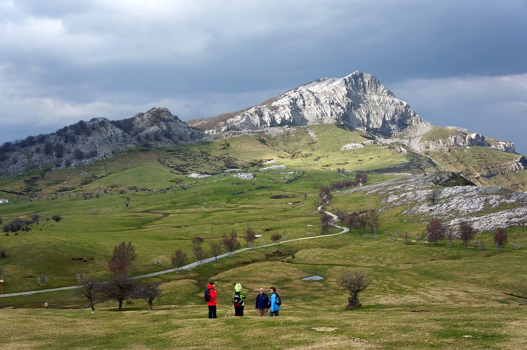 Wide view of Gorbea Natural Park with rugged limestone mountains, green pastures, scattered trees, and hikers under a cloudy sky.