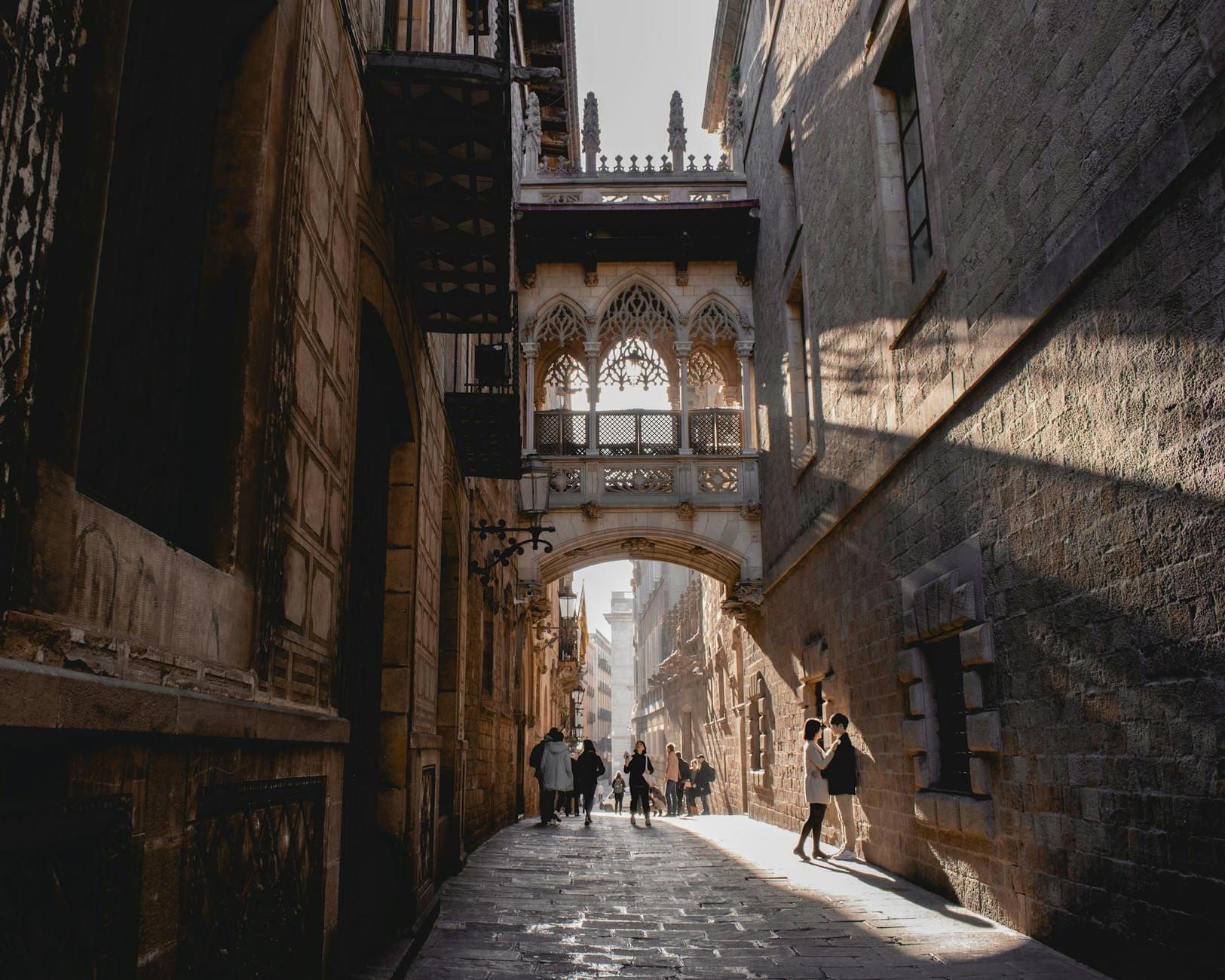 A narrow cobblestone street in Barcelona's Gothic Quarter with historic stone buildings and the iconic Pont del Bisbe bridge, sunlight streaming in, and people walking.
