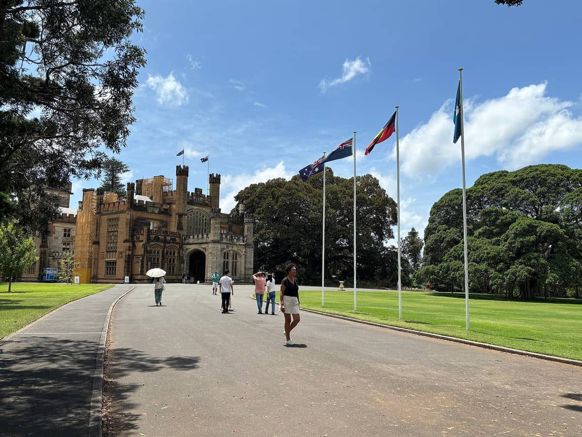 La storica Government House di Sydney con la sua architettura neogotica, circondata da rigogliosi giardini, diverse bandiere nazionali e visitatori che passeggiano sotto un cielo azzurro brillante.
