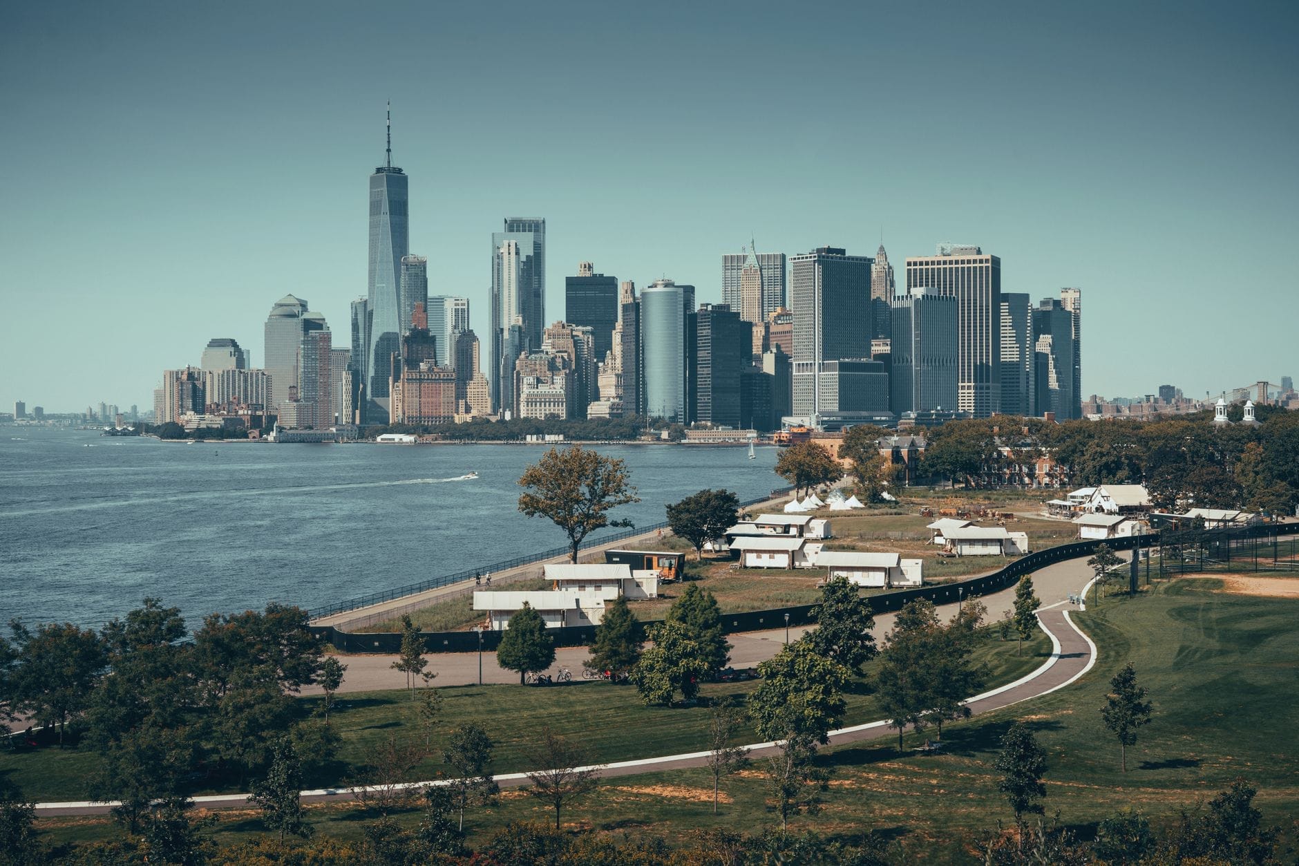 Governors Island in primo piano con spazi verdi e sentieri pedonali, lo skyline di Lower Manhattan che si staglia oltre il porto in una giornata limpida.