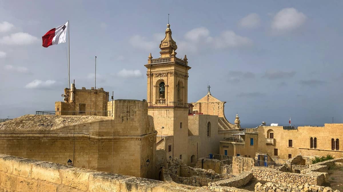 Widok na twierdzę Cittadella w Victorii, Gozo, z maltańską flagą, katedrą i średniowiecznymi kamiennymi murami pod zachmurzonym niebem.