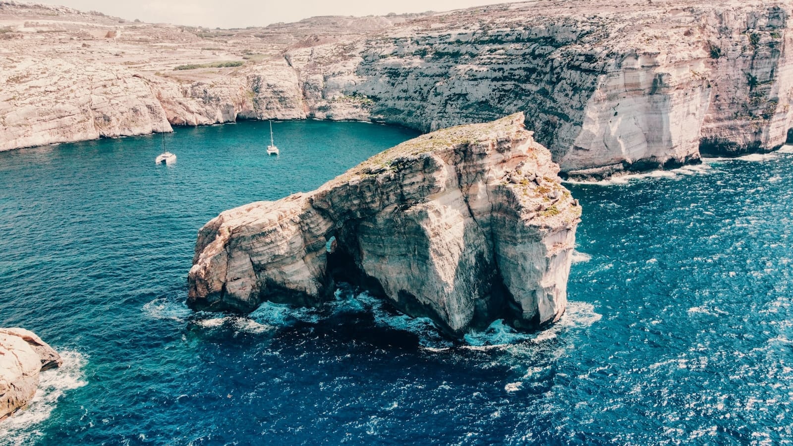 Falésias dramáticas e uma grande formação rochosa cercada por água turquesa em Gozo, Malta, vista de cima em um dia ensolarado.