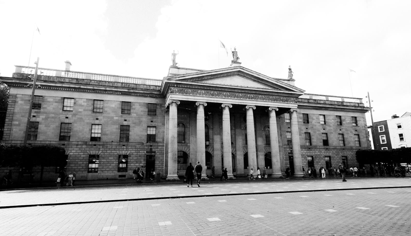 Ampia fotografia in bianco e nero del General Post Office di Dublino con le sue iconiche colonne e alcune persone che camminano davanti su O'Connell Street.