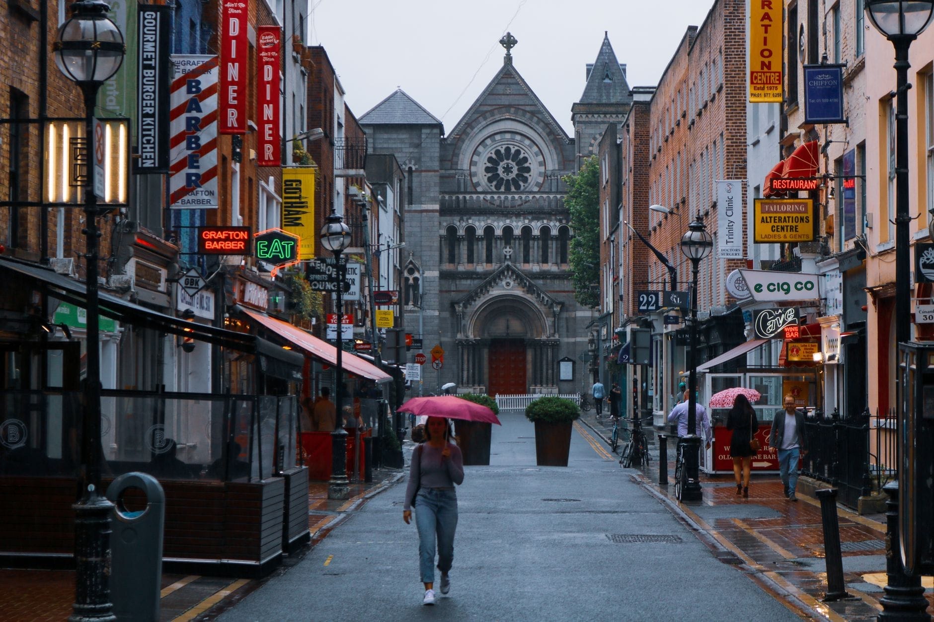Vista di Grafton Street a Dublino in un giorno di pioggia, con insegne di negozi colorate, pedoni e una chiesa storica in fondo.