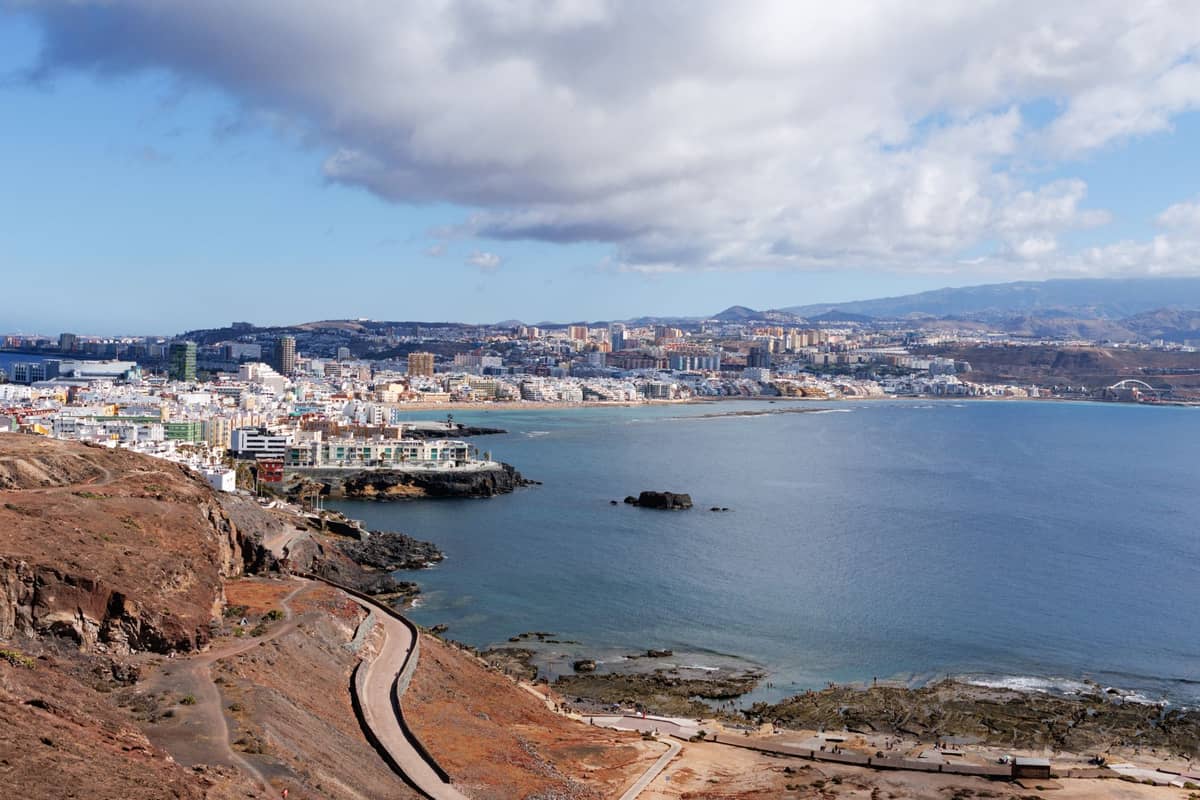 Wide landscape view of Las Palmas coastline and cityscape in Gran Canaria, with buildings stretching along the shore and mountains in the background under a partly cloudy sky.