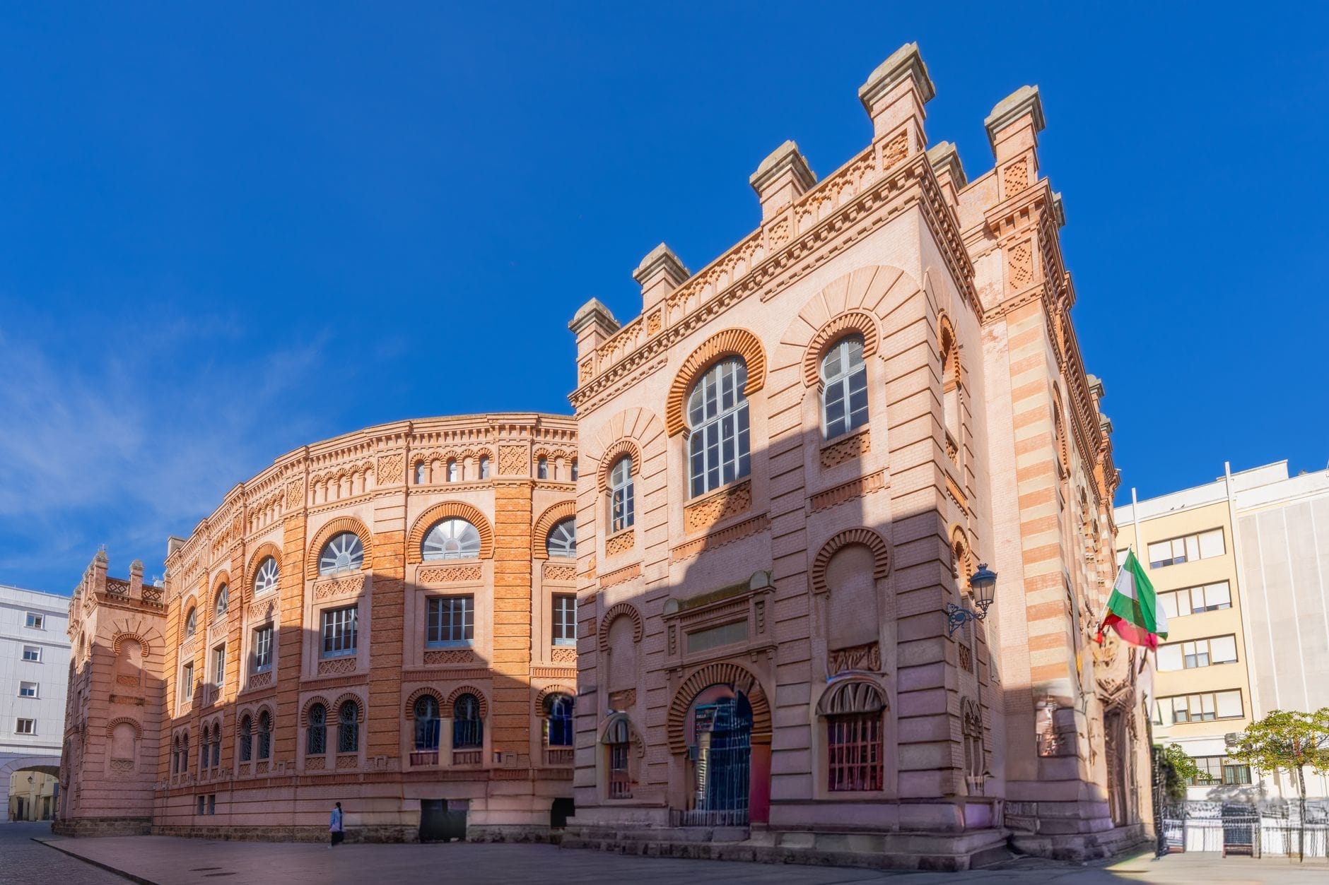 Vista frontale della spettacolare facciata in mattoni rossi neo-mudéjar del Gran Teatro Falla, sotto un cielo azzurro terso a Cádiz, Spagna, con luce solare intensa e piazza deserta.