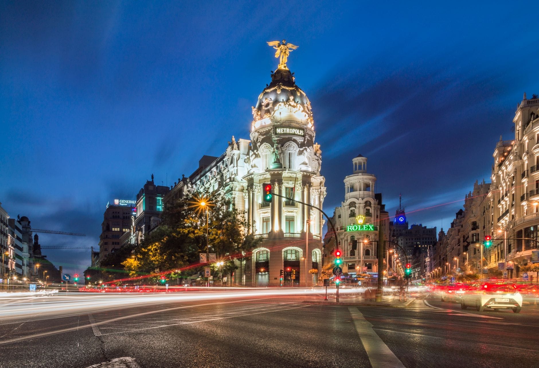 Vista notturna della Gran Vía di Madrid con l'illuminato Edificio Metropolis, il viale animato e le luci vibranti della città sotto un cielo blu intenso.