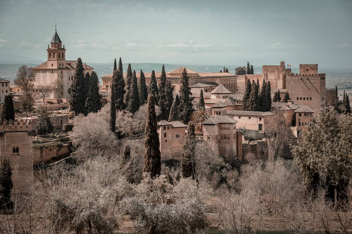 วิวพาโนรามาของกลุ่มอาคาร Alhambra พร้อมหอคอยอันเป็นสัญลักษณ์และพืชพรรณสีเขียวชอุ่ม มองเห็นจากระยะไกลใต้ท้องฟ้าแจ่มใส