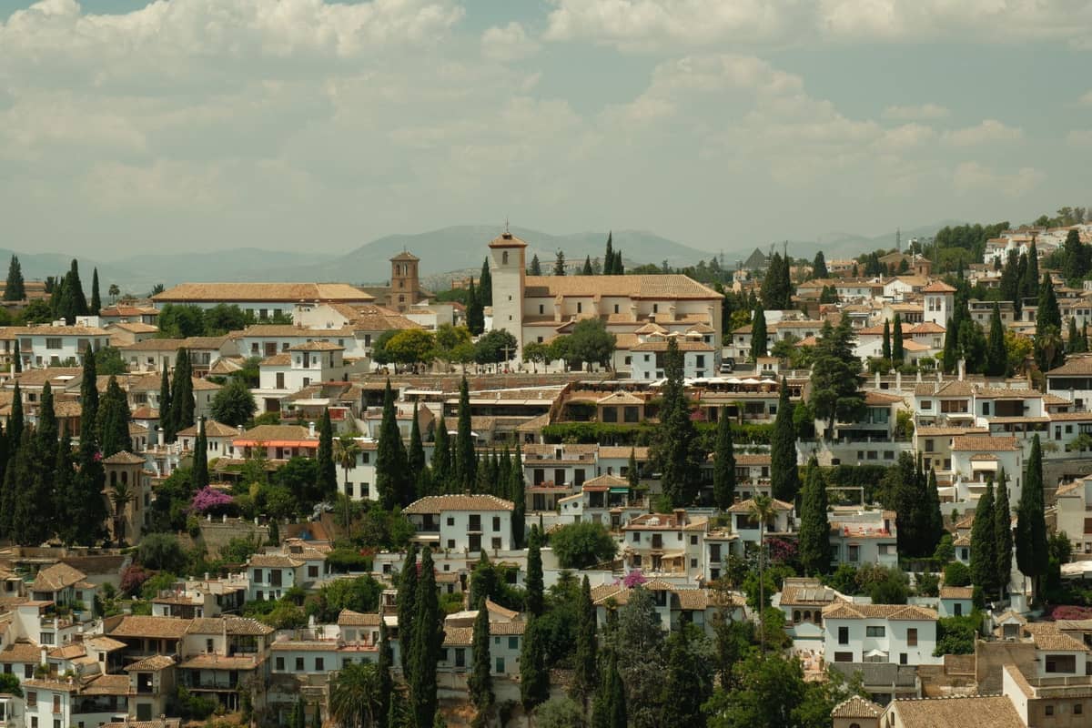 วิวพาโนรามาของย่าน Albaicín ในเมือง Granada พร้อมบ้านทาสีขาว ต้นไซเปรส และเนินเขาใต้ท้องฟ้ามีเมฆบางส่วน