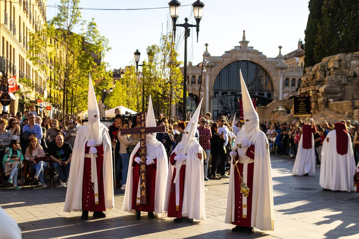 ขบวนแห่อีสเตอร์พร้อม nazarenos สวมชุดคลุมสีขาวและหมวกแหลม เดินขบวนผ่านจัตุรัสเมืองที่คึกคักเต็มไปด้วยผู้คนและอาคารประวัติศาสตร์