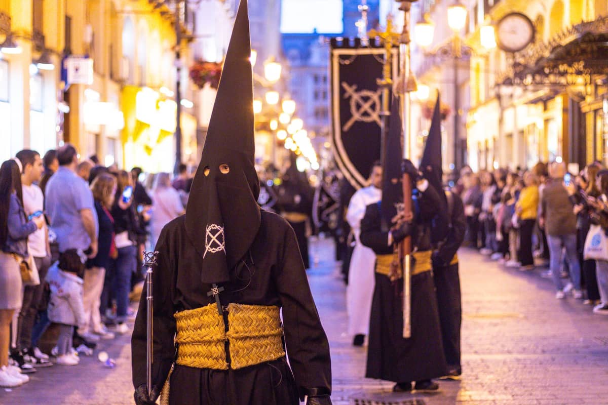 ขบวน Nazarenos สวมชุดสีดำและหมวกปลายแหลมเดินในขบวน Semana Santa ผ่านถนนสว่างไสวของกรานาดา รายล้อมด้วยฝูงชนที่มาดูขบวนแห่สัปดาห์ศักดิ์สิทธิ์