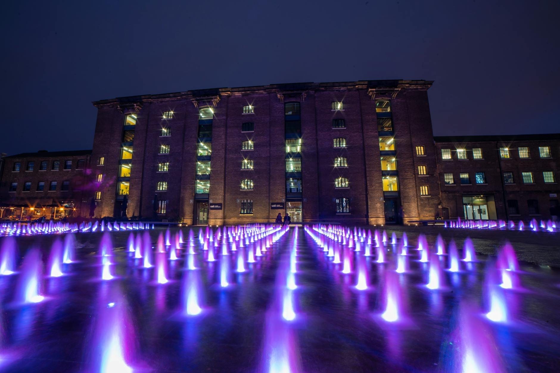 Vista notturna delle fontane illuminate di Granary Square con luci viola davanti allo storico Granary Building a King's Cross, Londra.