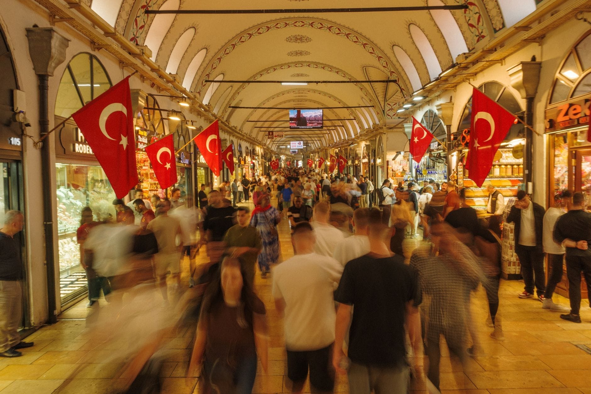 Interno animato del Grande Bazar di Istanbul, con folle di visitatori, bandiere turche, soffitti ad arco e negozi variopinti che si affacciano su un ampio corridoio.
