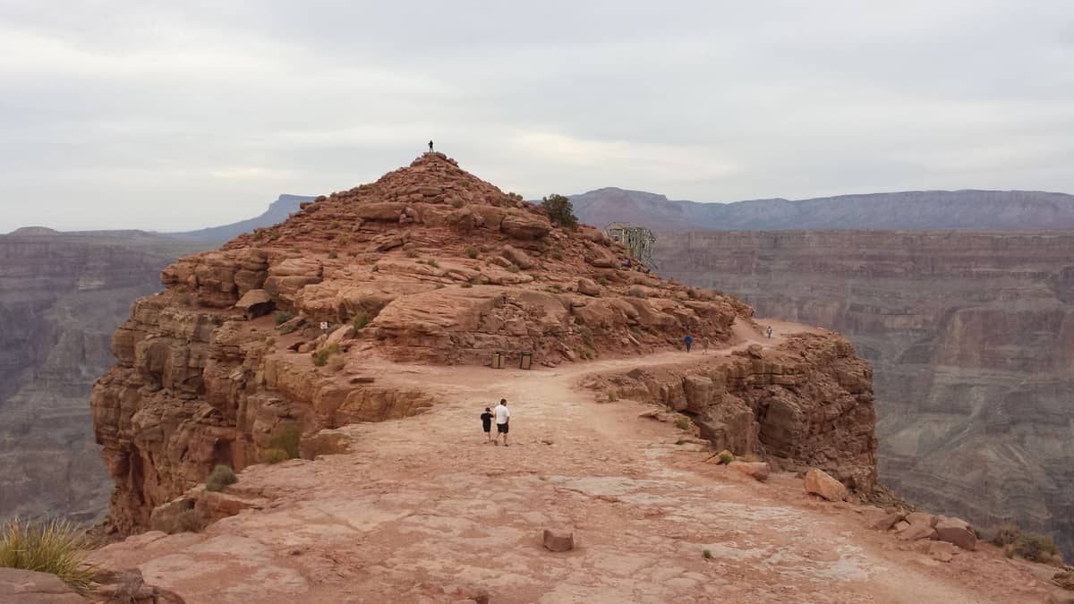 Visitantes caminham pelo planalto rochoso do Grand Canyon West Rim, em terras Hualapai, com o imenso canyon se espalhando ao fundo sob um céu nublado.