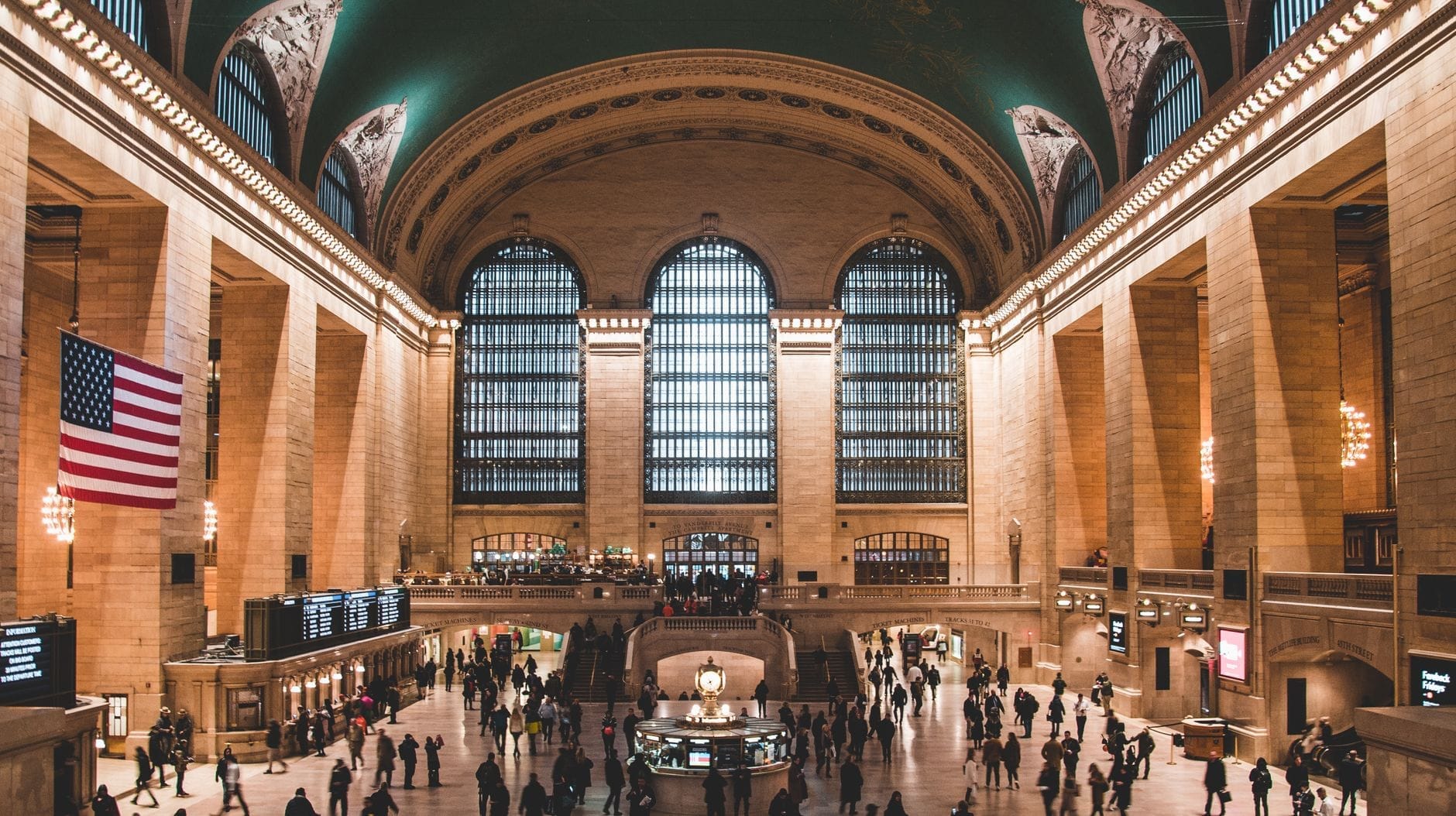 Vista grandangolare della sala principale della Grand Central Terminal con le sue iconiche finestre ad arco, la bandiera americana e la folla sotto il soffitto verde con le costellazioni dorate.