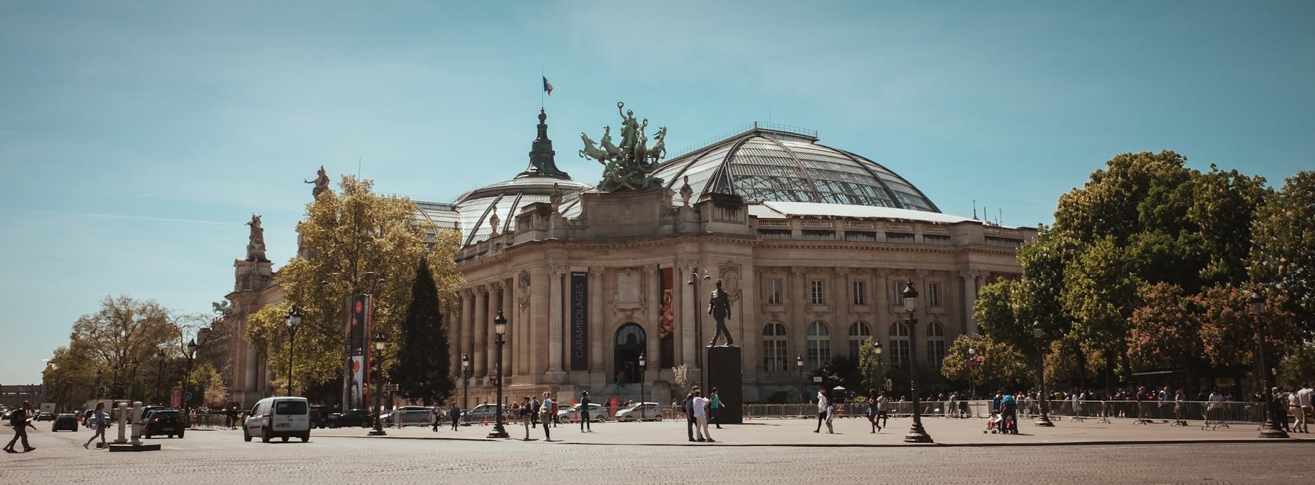 Vista grandangolare del Grand Palais a Parigi sotto un cielo azzurro limpido, con la cupola in vetro e l'architettura ornata in primo piano tra persone e auto.