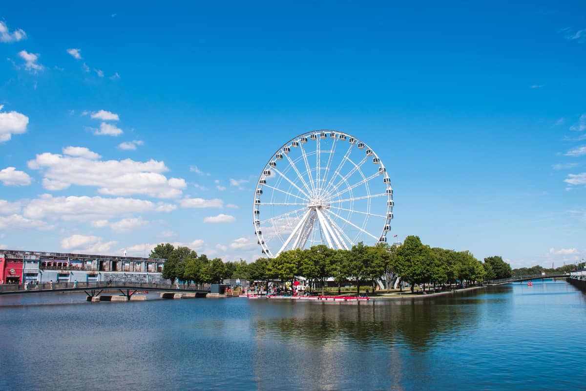 Das Riesenrad La Grande Roue de Montréal ragt über die Uferpromenade des Alten Hafens hinaus, mit blauem Himmel, Bäumen und Wasser im Vordergrund.