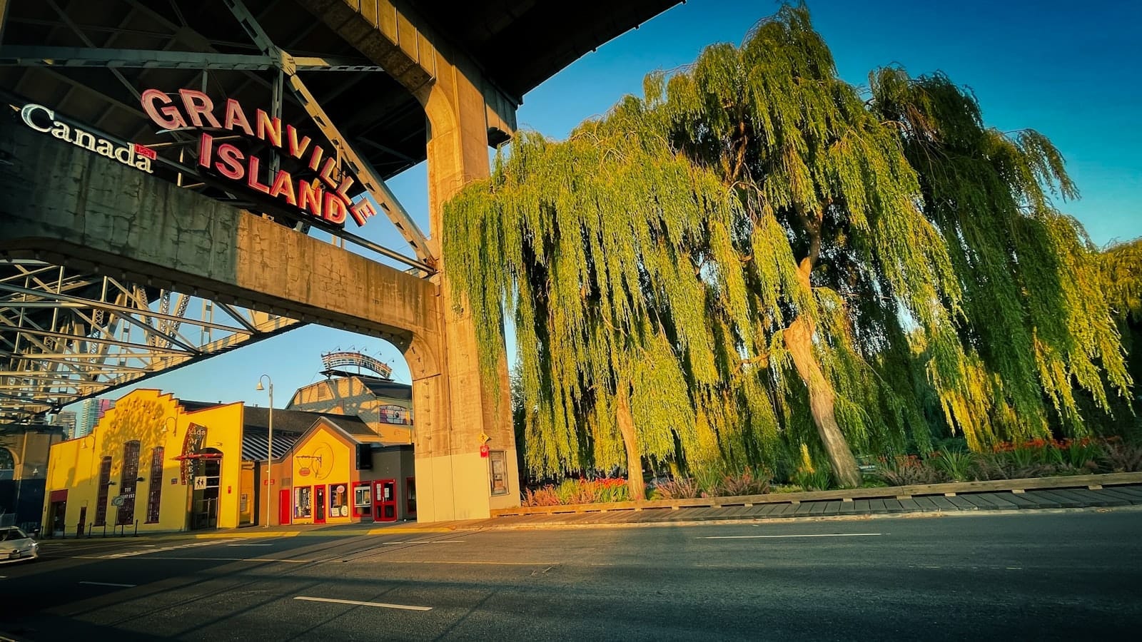 ป้ายทางเข้า Granville Island ใต้สะพาน ตึกสีเหลืองต้นไม้ใบเขียว แสงแดดสีทองสดใส