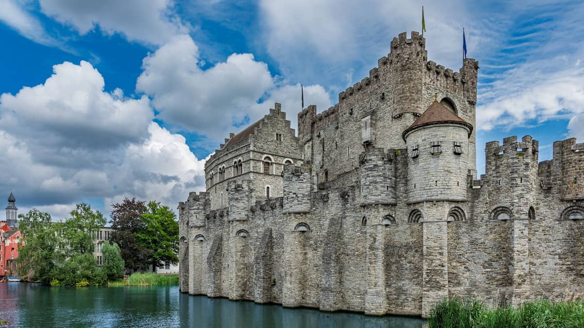 Wide landscape view of Gravensteen Castle in Ghent with its stone walls, towers, and moat under a partly cloudy sky.