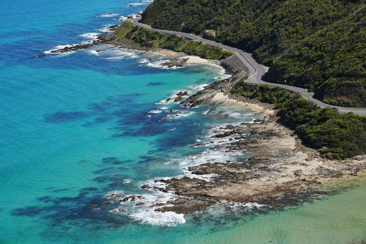 Vista aérea de la Great Ocean Road serpenteando a lo largo de la accidentada costa de Victoria con aguas turquesas, olas y exuberante vegetación verde en los acantilados.