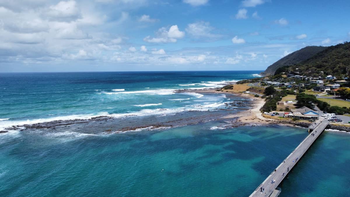 Luftaufnahme der Küste von Lorne an der Great Ocean Road mit einem Pier, blauem Ozean und teilweise bewölktem Sommerhimmel.