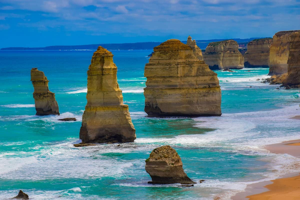 Vista clara y luminosa de las formaciones de piedra caliza de los Doce Apóstoles emergiendo del océano turquesa a lo largo de la Great Ocean Road, con olas rompiendo en la playa de arena.