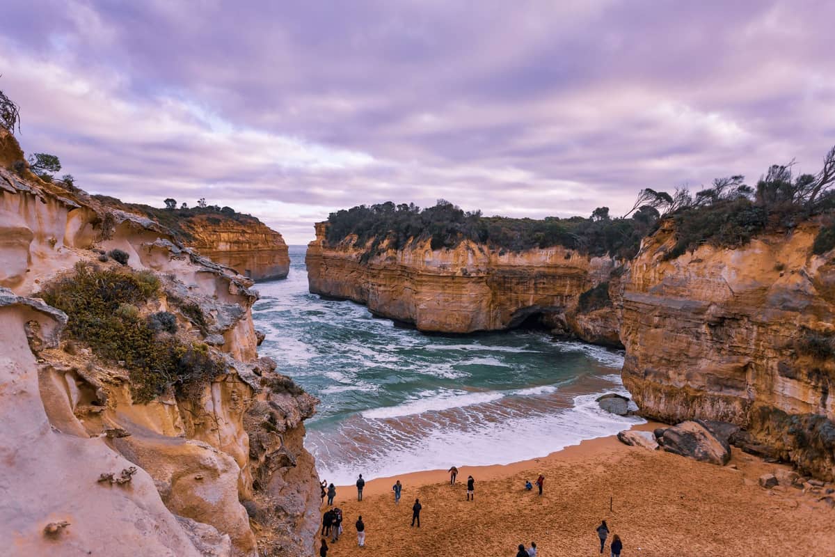 Blick auf die Loch Ard Gorge mit Sandstrand, Spaziergängern und beeindruckenden Klippen auf beiden Seiten unter einem bewölkten Himmel.