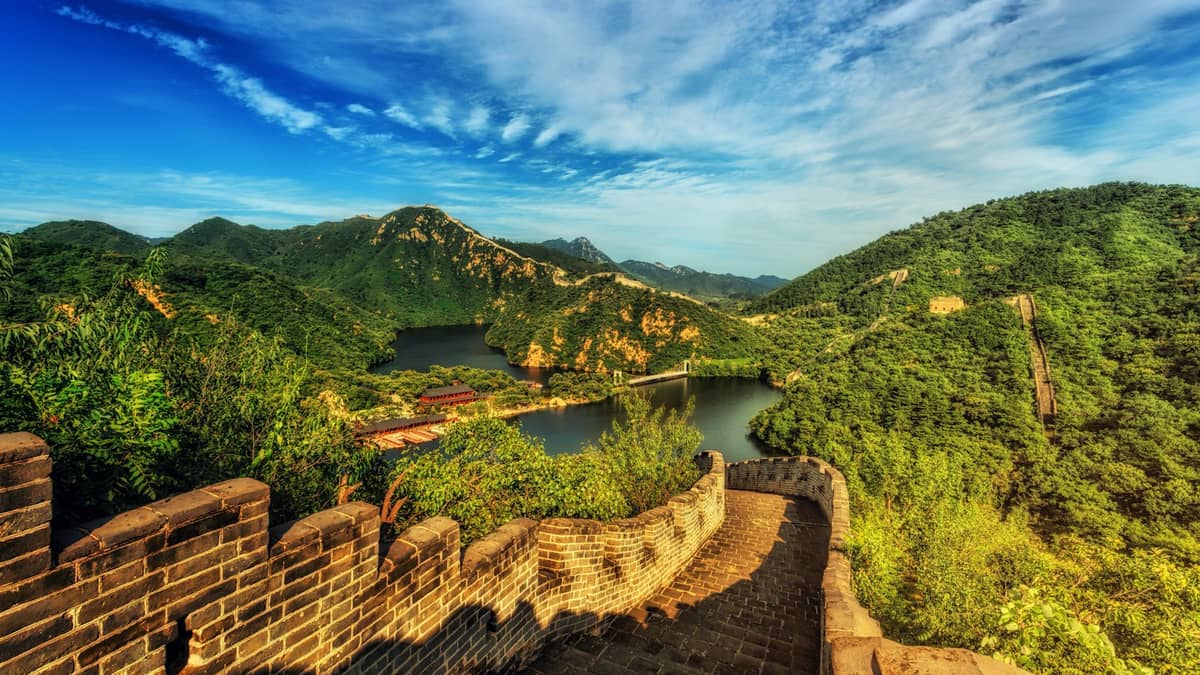 Die Chinesische Mauer schlängelt sich über üppig grüne Berge, im Hintergrund ein Fluss und blauer Himmel, aufgenommen bei Tageslicht.