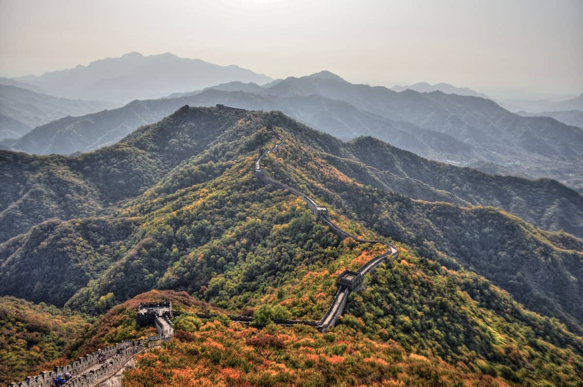 Vista panorámica de la Gran Muralla China atravesando colinas de colores otoñales bajo un cielo brumoso, con escasas multitudes y follaje pintoresco.