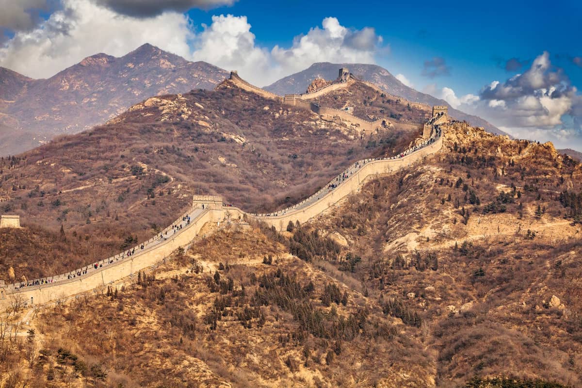 Vista panorámica del paisaje de la Gran Muralla China serpenteando sobre montañas escarpadas bajo un cielo azul brillante con nubes dispersas.