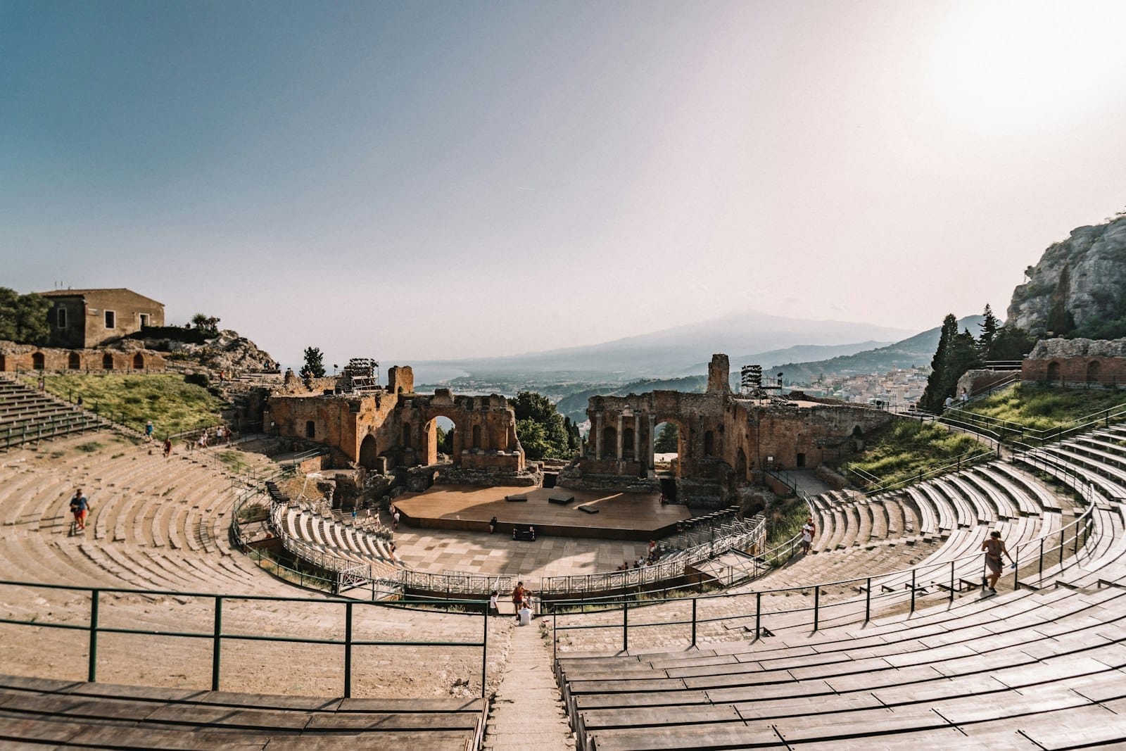 Weitwinkelaufnahme des antiken griechischen Theaters von Taormina mit steinernen Sitzreihen, beeindruckenden Ruinen, dem Ätna und dem Ionischen Meer in der dunstigen Ferne unter klarem Himmel.