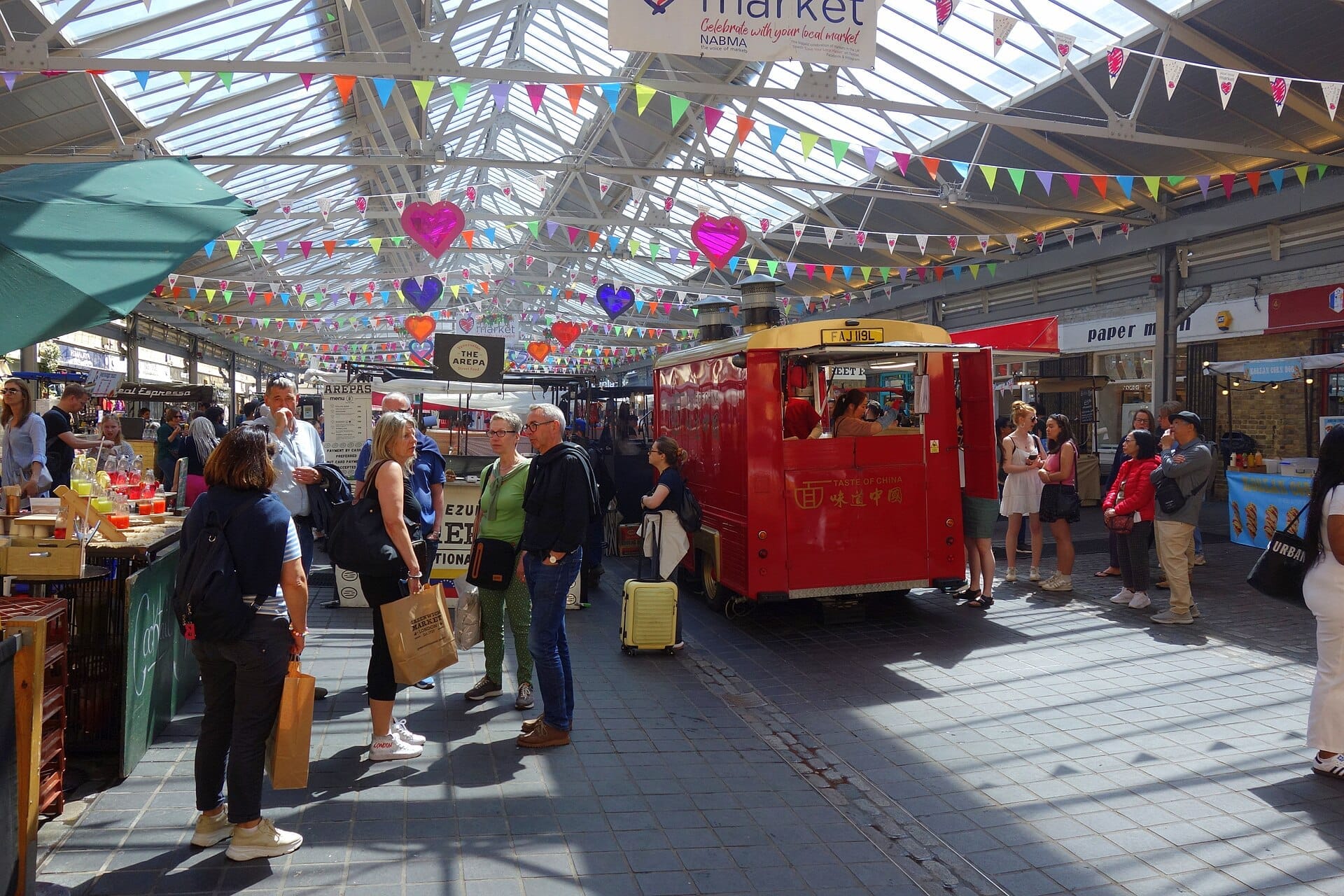 I visitatori esplorano il vivace mercato al coperto di Greenwich Market, con festoni colorati, bancarelle di cibo e un vintage food truck rosso sotto un tetto di vetro.