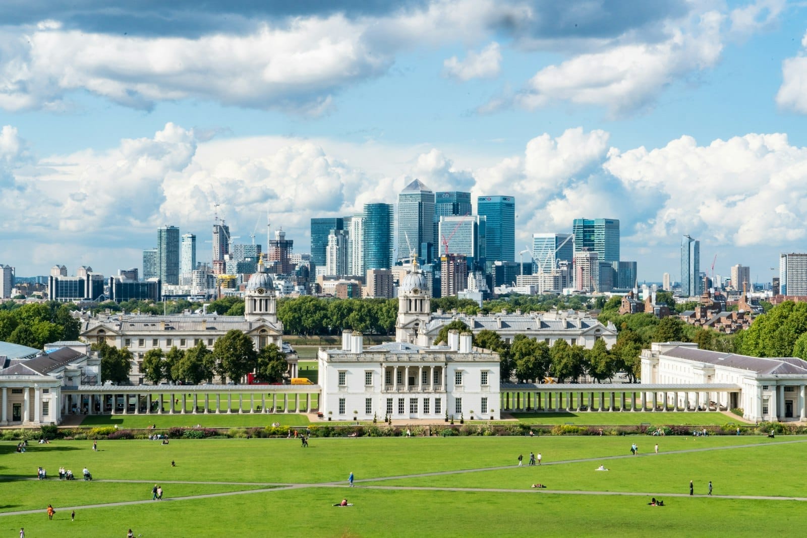 Vista dei prati verdi di Greenwich Park e dei suoi grandi edifici storici con il moderno skyline di Canary Wharf a Londra sotto un cielo parzialmente nuvoloso.