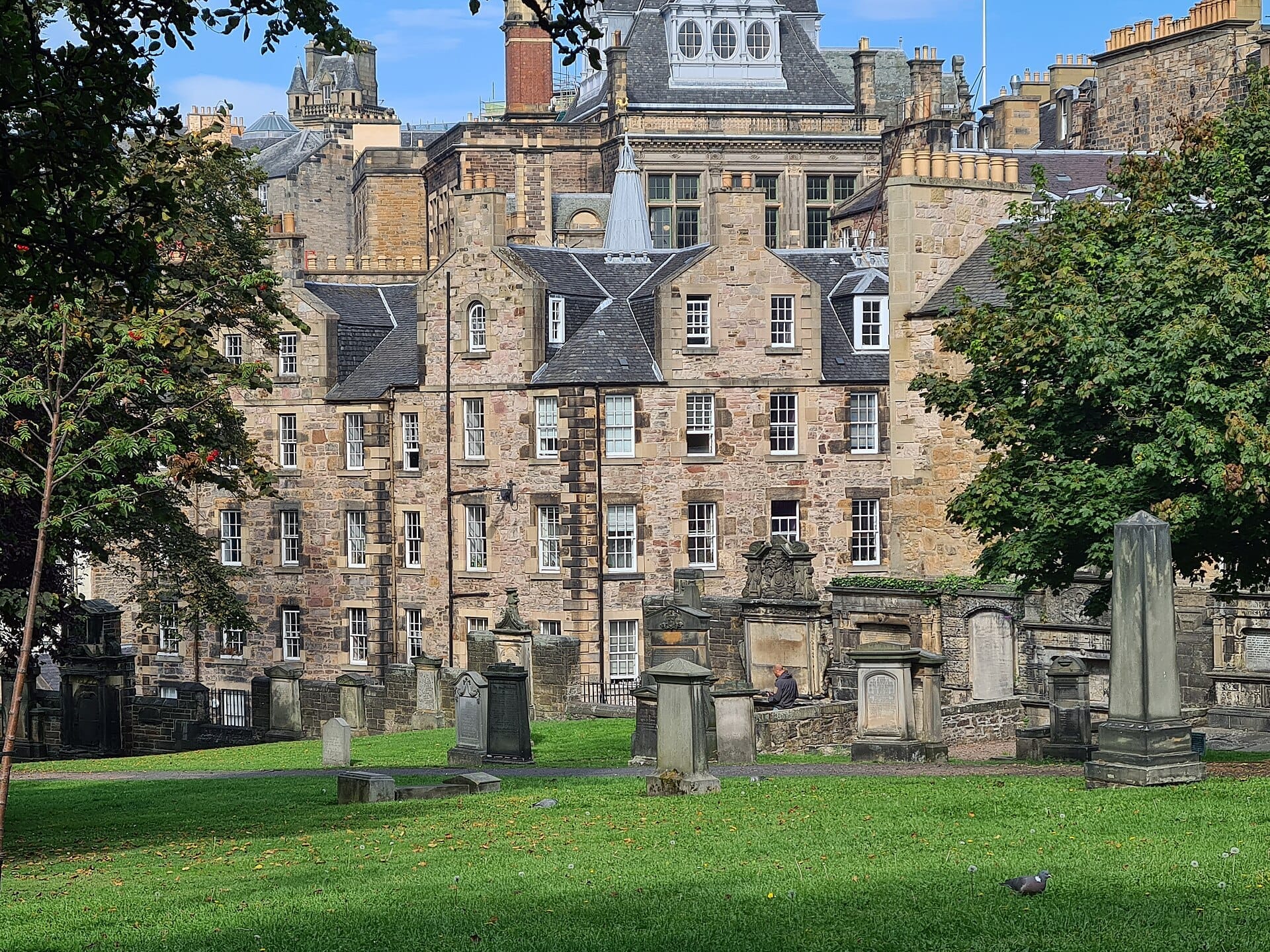 Reihen verwitterter Grabsteine und üppig grünes Gras im Greyfriars Kirkyard, vor historischen Steingebäuden unter klarem Himmel.