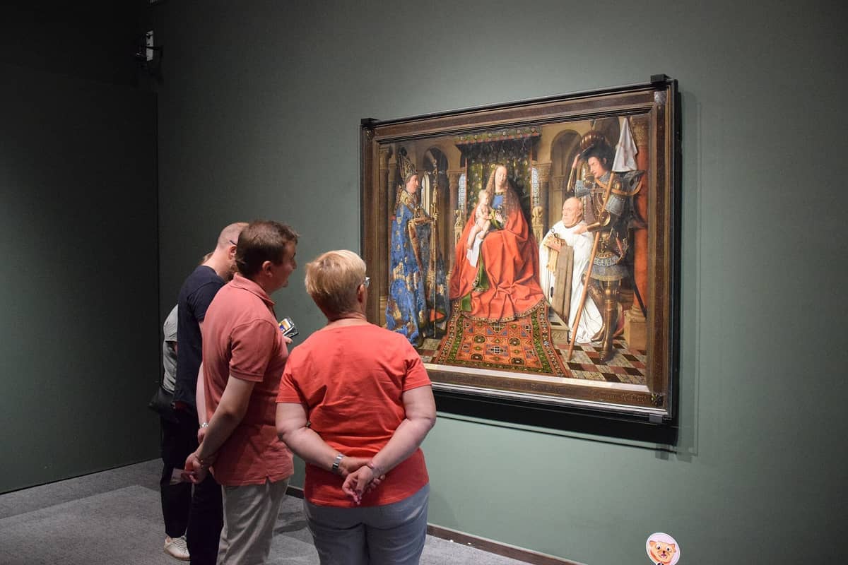 Visitors at the Groeningemuseum in Bruges closely examine an ornate Flemish masterpiece displayed on a green wall under gallery lighting.