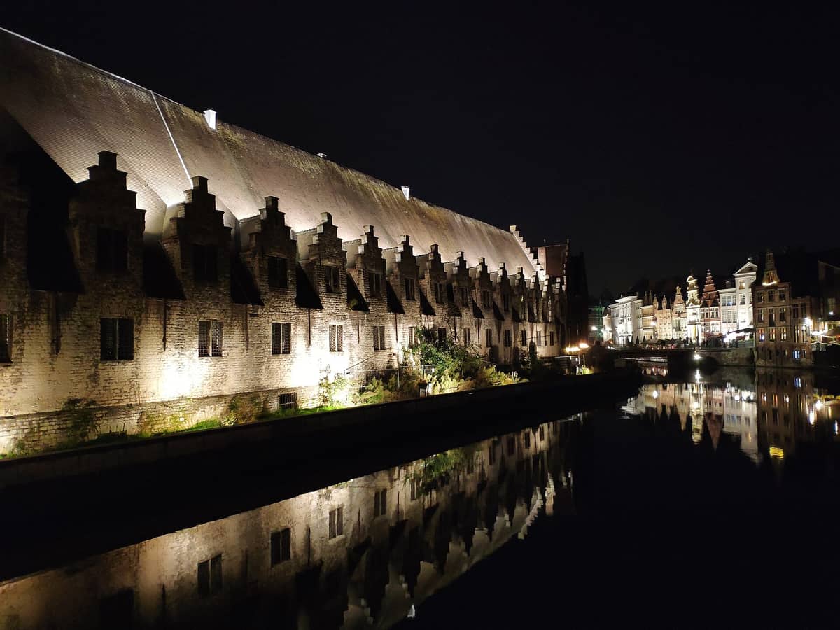 Night view of the Groot Vleeshuis in Ghent, its long medieval stone structure illuminated and reflected in the adjacent canal with historic buildings nearby.