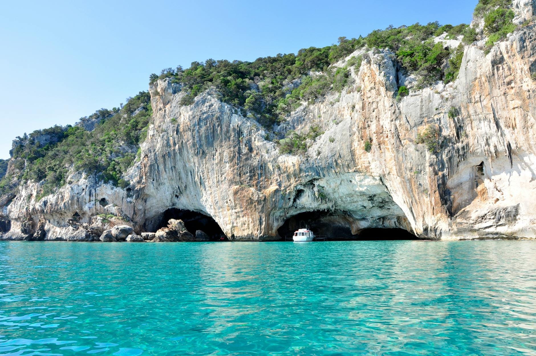 Blick auf die Eingänge der Meereshöhle Grotte del Bue Marino in Kalksteinklippen mit türkisblauem Wasser und einem Boot vor der Höhle unter klarem blauen Himmel.