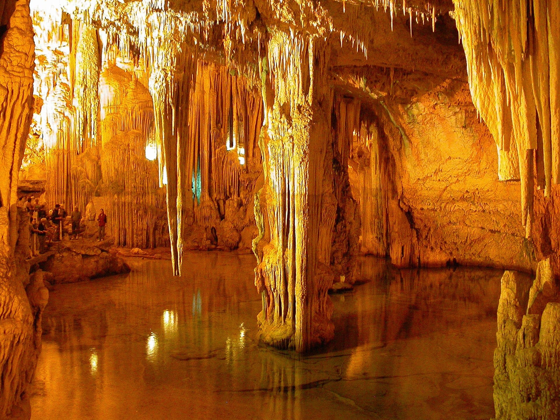 Besucher gehen entlang eines Weges in den Grotte di Nettuno, umgeben von beeindruckenden Stalaktiten, Stalagmiten und einem spiegelnden unterirdischen See unter warmem Licht.